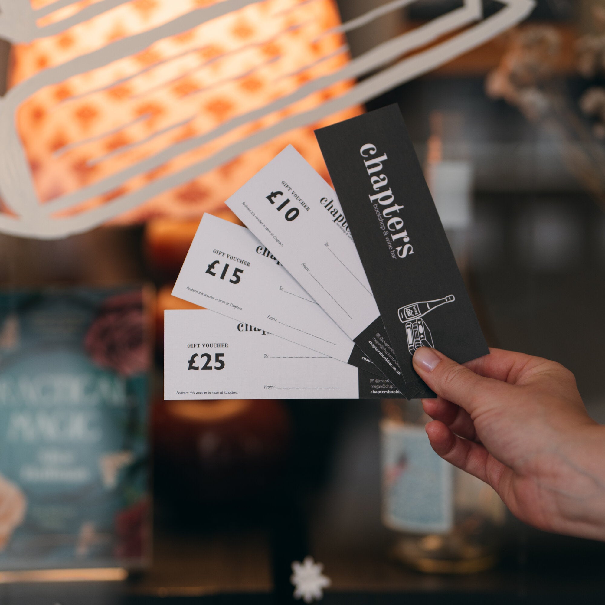 A hand holding a fan of Chapters gift vouchers for £10, £15 and £25 with a branded card, in the shop window